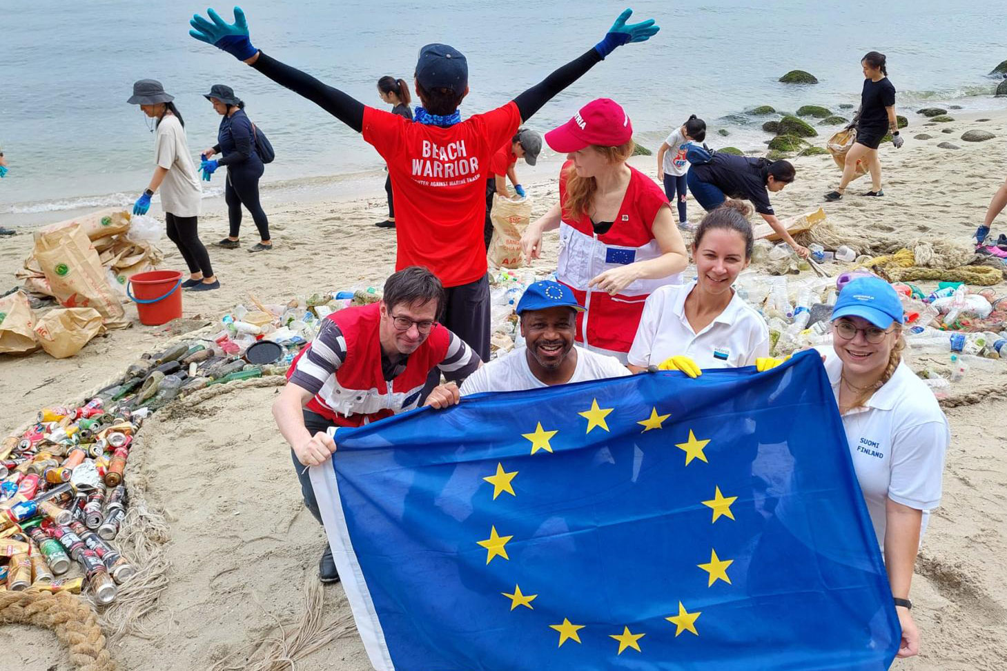 2024 EUBeachCleanup, Team Europe cleanup with Singapore Beach Warriors. People on a beach picking up rubbish and 4 people holding an EU flag