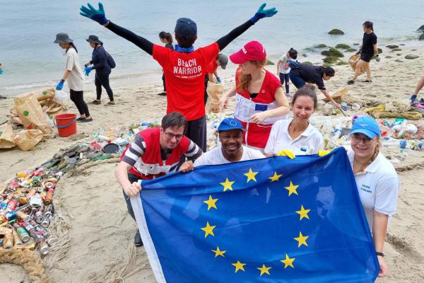 2024 EUBeachCleanup, Team Europe cleanup with Singapore Beach Warriors. People on a beach picking up rubbish and 4 people holding an EU flag